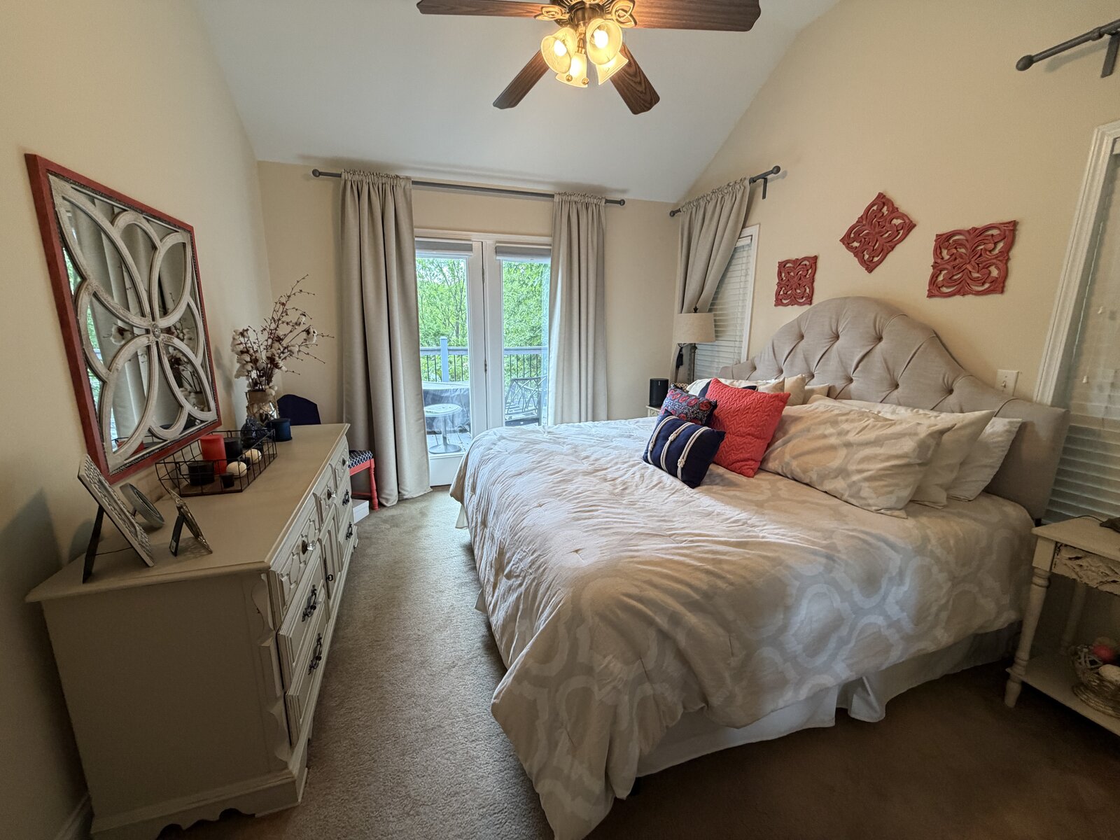Primary bedroom after renovation — a tufted upholstered headboard, soft neutral bedding, and coral accents by the balcony doors