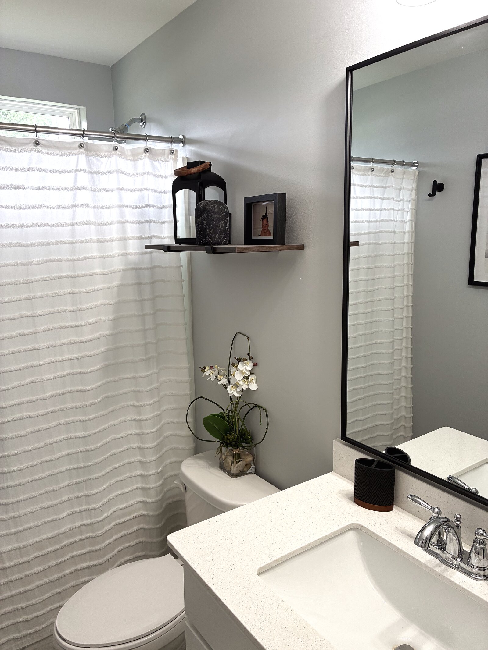 Guest bath — grey walls with a black-framed mirror, textured white shower curtain, and a floating shelf styled with a black lantern