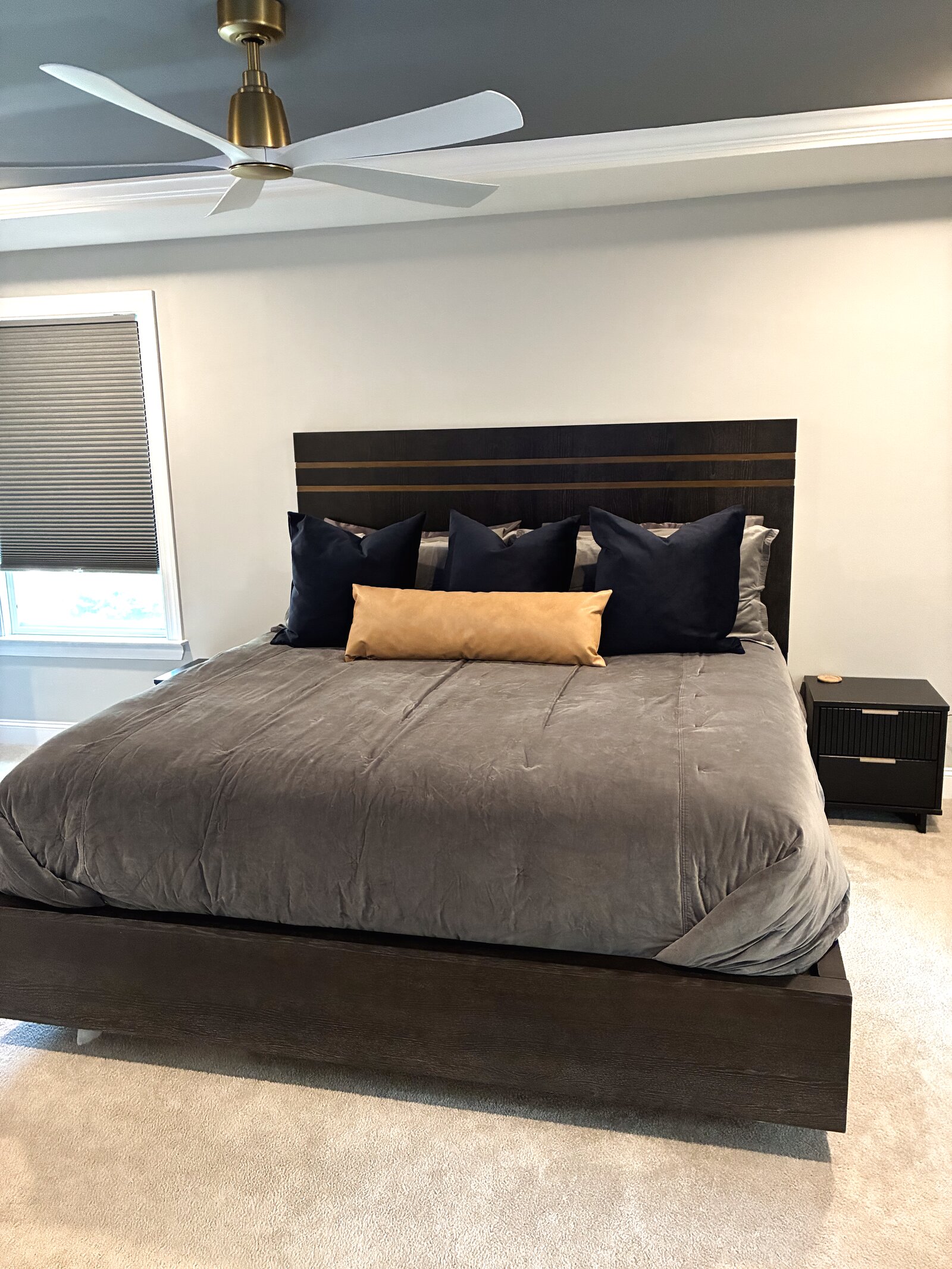 Primary bedroom — dark platform bed with a slatted brass-inlay headboard, grey velvet duvet, navy pillows, and a camel-leather lumbar