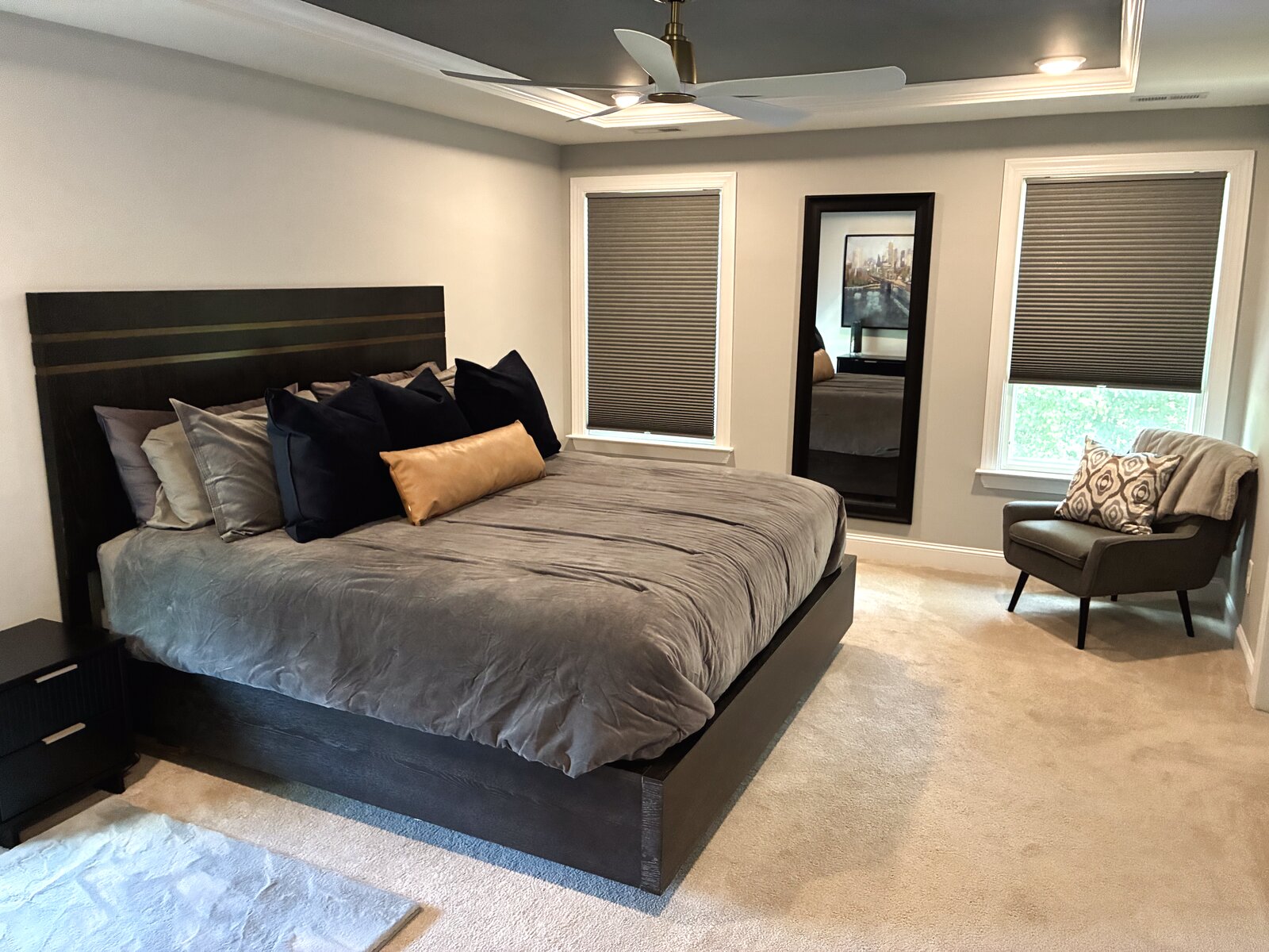 Moody primary bedroom with a dark platform bed and slatted headboard, grey velvet bedding, navy and camel-leather pillows, a black dresser, and a tray ceiling