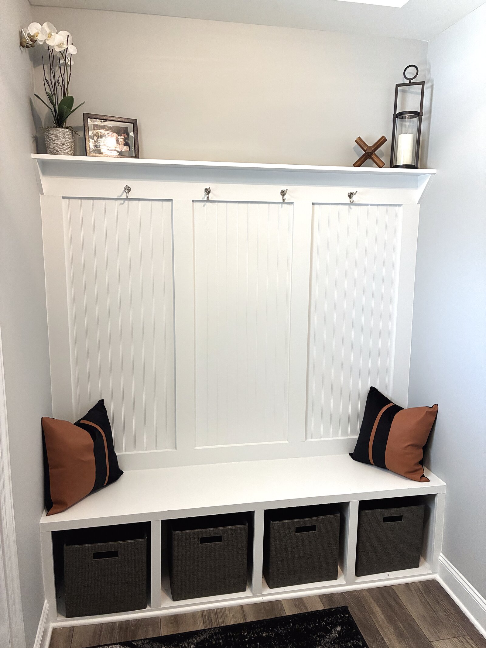 Entry drop zone — white built-in bench with cubby baskets, beadboard hooks, a display shelf, and camel-and-black striped pillows