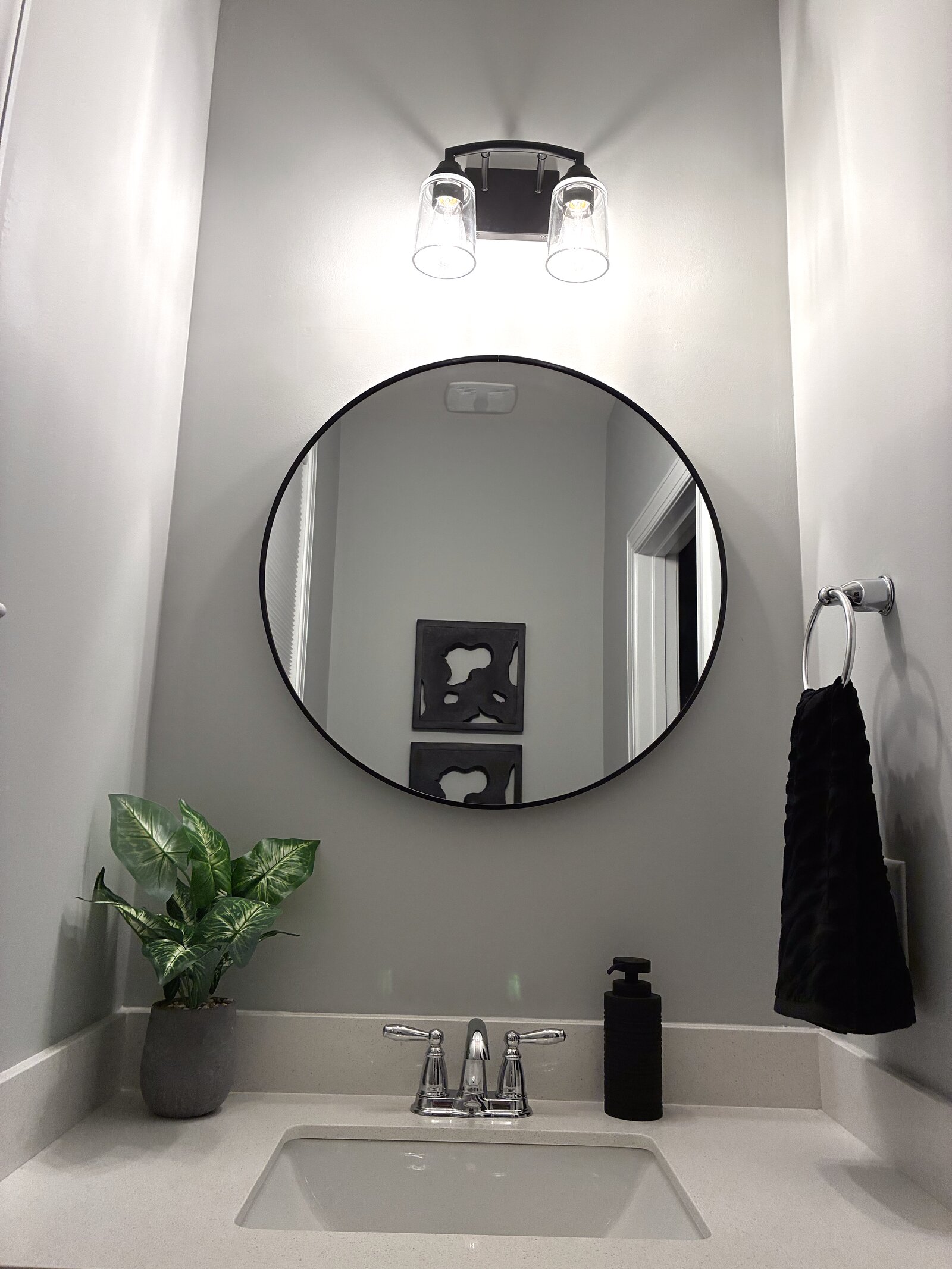 Powder room — round black-framed mirror under a two-globe fixture, chrome faucet, black accents, and a potted plant on a white vanity